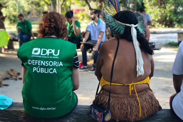 Fotografia colorida de indígena sentada ao lado de mulher com colete verde