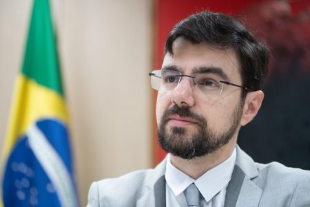 Foto colorida de secretário Guilherme Mello, de terno e óculos em frente a bandeira do Brasil - Metrópoles