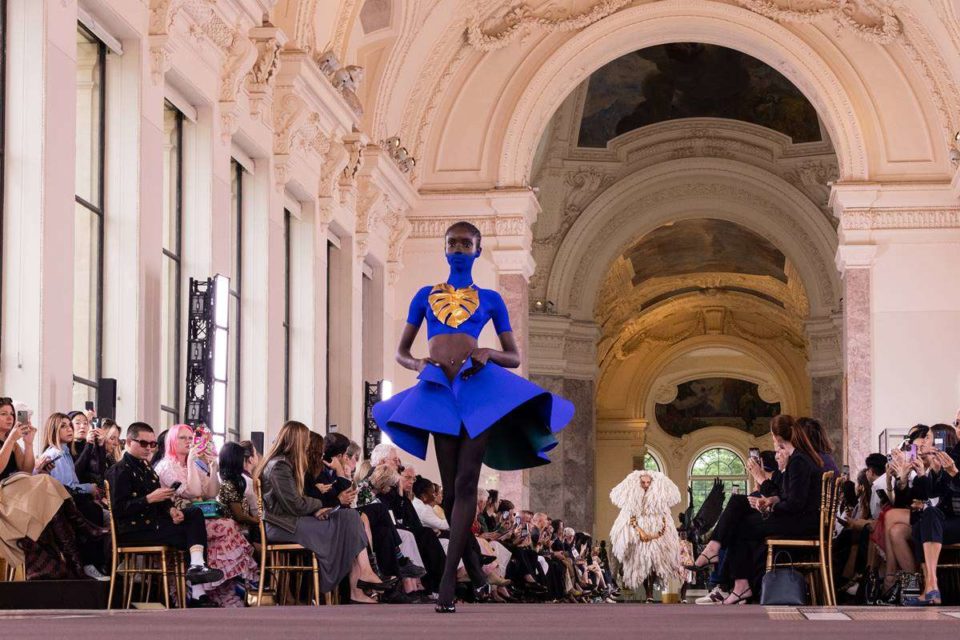 Em desfile, ao centro, modelo negra caminha pela passarela, em meio ao público posicionado nas laterais em cadeiras. Ela tem parte do pescoço e do rosto pintados de azul, na mesma tonalidade do look com saia e cropped, que tem uma folha dourada - Metrópoles