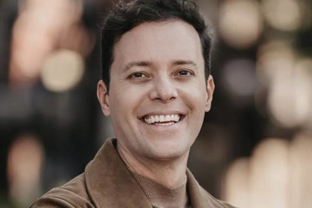 Foto colorida de André Valadão. Ele é branco, tem cabelo preto e sorri para a foto. Na imagem, ele utiliza roupas brancas - Metrópoles