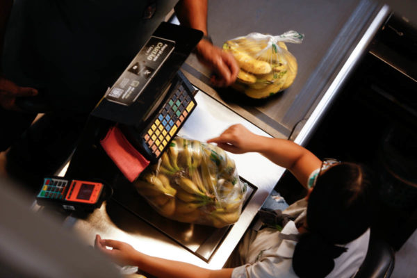 Imagem colorida mostra mulher pesando banana no caixa de mercado - Metrópoles