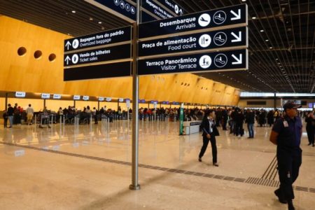 Placas em aeroporto com pessageiros passando ao fundo - Metrópoles