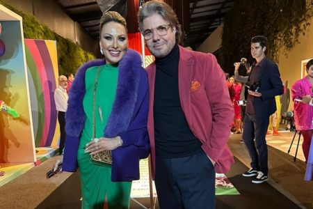 João Kleber está de calça jeans, óculos de grau vermelho, camisa preta e terno vermelho, enquanto Mara Ferraz escolheu um casaco azul, um vestido verde e uma bolsa beje - Metrópoles