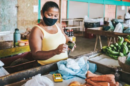 Mulher negra, de máscara, observa maquininha de cartão enquanto segura o celular em um mercado - Metrópoles