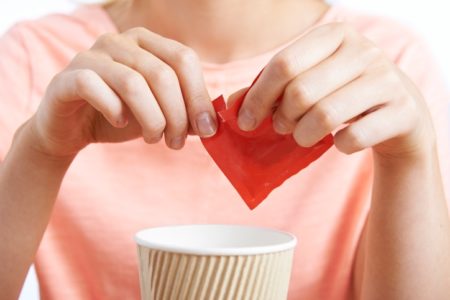 Foto de mulher que abre saco de adoçante aspartame diante de um copo descartável de bebida - Metrópoles