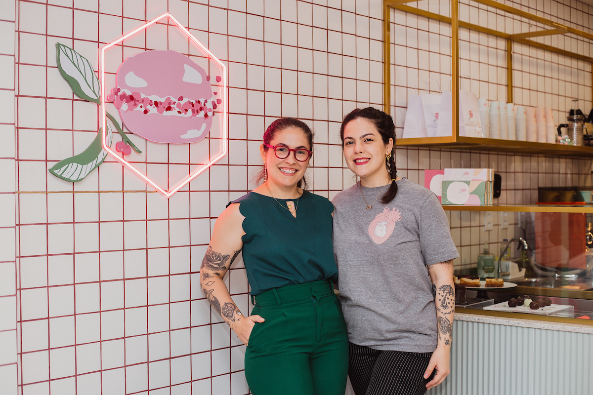 Foto colorida de duas mulheres brancas e abraçadas próximo a uma imagem de um macaron gigante pintada em uma parede. As mulheres estão abraçadas - Metrópoles