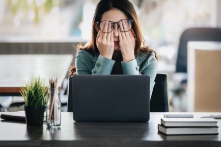 Foto mostra mulher pressionando os olhos diante de um computador em um ambiente laboral - Metrópoles