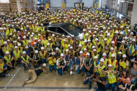 Imagem colorida de funcionários da Tesla, com coletes amarelos e capacetes brancos, posando ao lado do Cybertruck