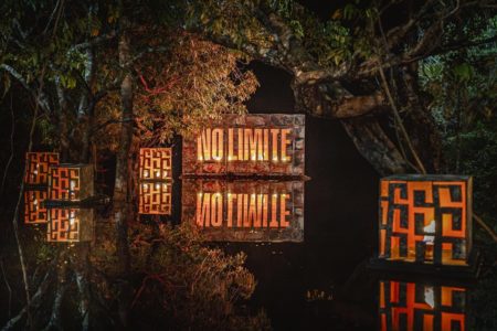 Foto colorida de No Limite - Amazônia. A foto mostra placas com a logo do No Limite, em meio à arvores e um lago - Metrópoles