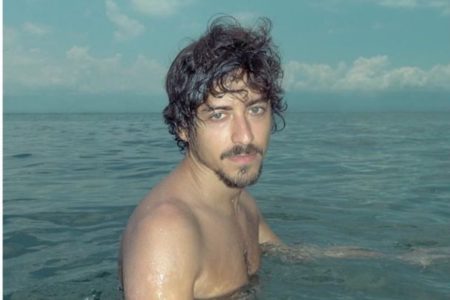 Foto colorida do ator Jesuíta Barbosa. Ele está sem camisa, dentro do mar, tem cabelo preto, barba preta e é branco - Metrópoles