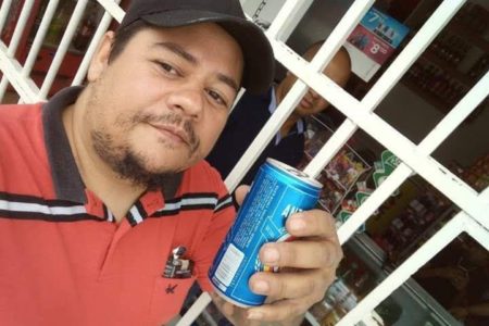 Homem pardo de cabelos negros segura lata de cerveja em frente a portão branco. Ele veste boné e uma camisa polo vermelha com listras pretas