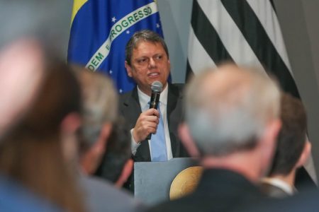 Homem branco de cabelo grisalho segura microfone em frente às bandeiras do Brasil e de São Paulo