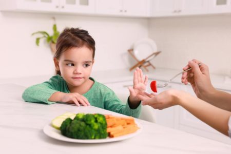 Na foto, mulher alimentando sua filha na cozinha e a menina se recusando a comer legumes - Metrópoles