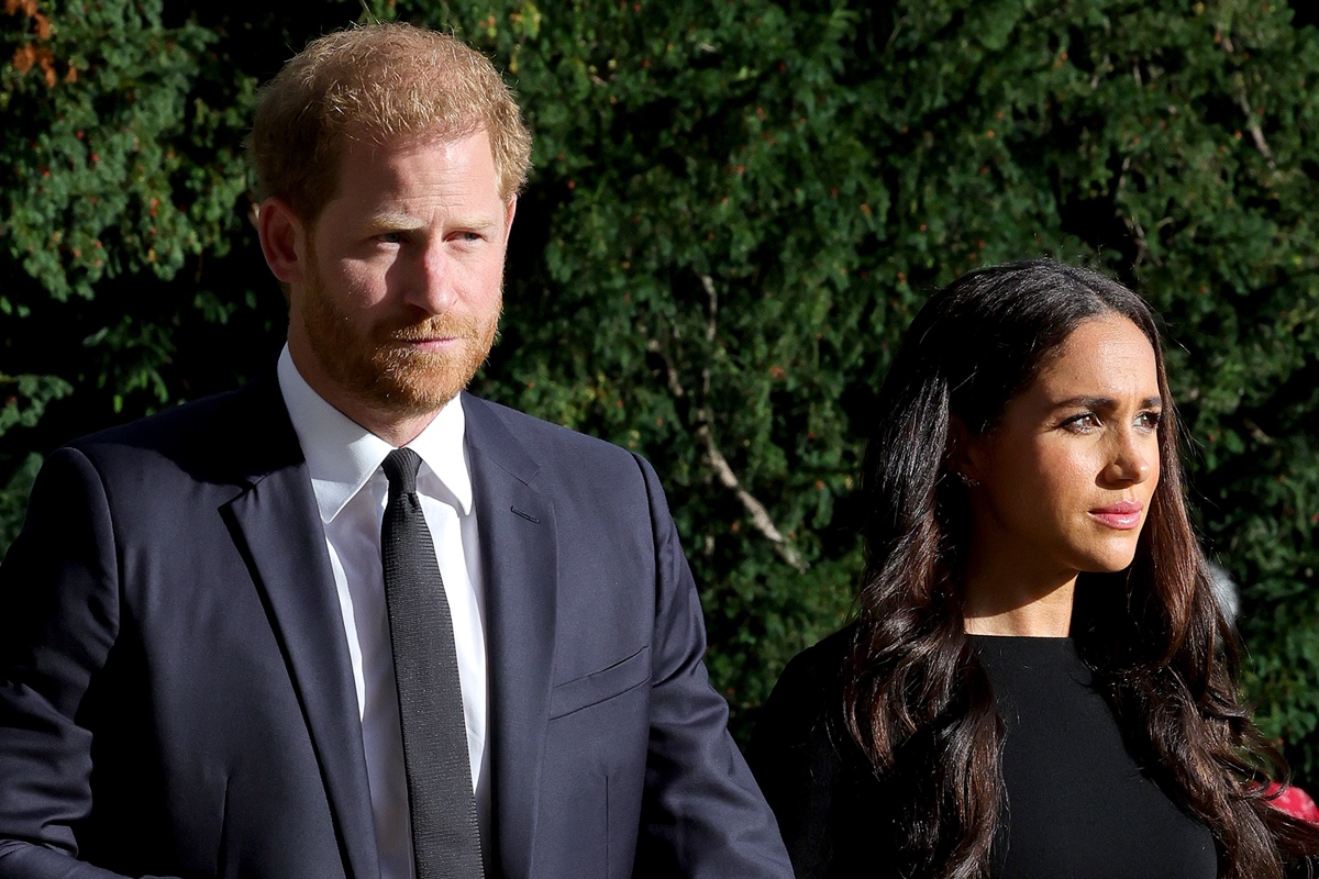 Foto colorida. Harry e Meghan são um casal e estão de mãos dadas. Ele é um homem branco, cabelos ruivos e está usando um terno azul escuro. Ela é uma mulher negra, cabelos longos pretos e liso. Está usando um vestido todo preto - Metrópoles