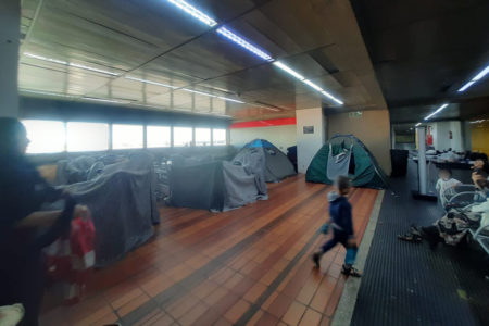 Imagem colorida mostra barracas montadas em corredor do aeroporto de guarulhos. Duas crianças estão na imagem, uma de costas, andando, e a outra olhando para a primeira. Uma mulher de hijab observa a cena na lateral - Metrópoles