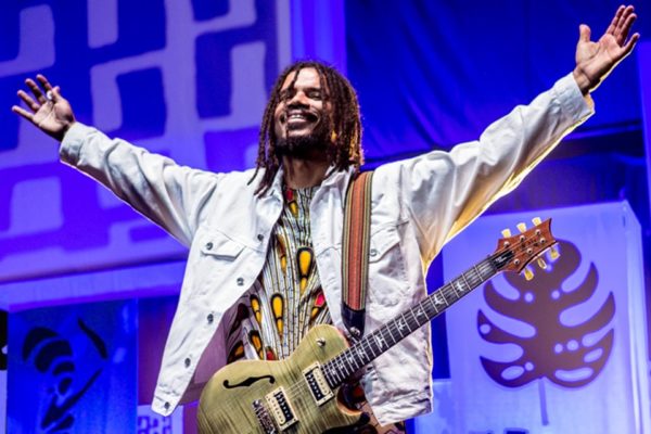 Alexandre, vocalista do Natiruts, comemora show em Buenos Aires, na Argentina. Ele está com uma jaqueta branca, camisa colorida e uma guitarra - Metrópoles