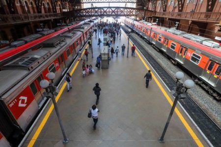 Imagem colorida mostra pessoas andando em passarela da estação luz. Dois trens estão parados de cada lado da plataforma. metrópoles