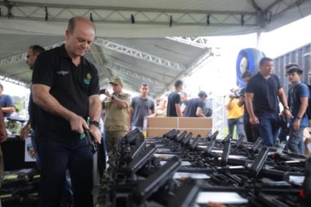 Imagem colorida de homem com arma na mão em frente de mesa com armas apreendidas - Metrópoles