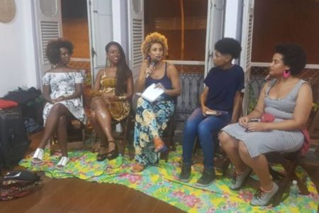 imagem colorida mostra cinco mulheres negras sentadas uma ao lado da outra. a terceira é a vereadora marielle franco, que segura papeis e fala com microfone nas mãos