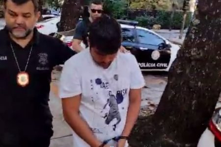 Torcedor do Flamengo de camiseta branca e cabeça baixa é conduzido por dois policiais até uma delegacia do Rio de Janeiro