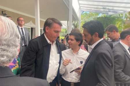 Imagem colorida com Bolsonaro, homem branco de camisa branca e jaqueta preta, um garoto e Ricardo Nunes, de lado, de terno cinza, em primeiro plano, com pessoas ao fundo, na área externa de uma casa. O garoto mostra a tela de seu celular para os outros dois - Metrópoles