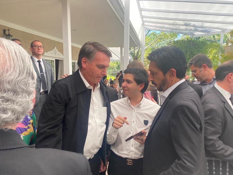 Imagem colorida com Bolsonaro, homem branco de camisa branca e jaqueta preta, um garoto e Ricardo Nunes, de lado, de terno cinza, em primeiro plano, com pessoas ao fundo, na área externa de uma casa. O garoto mostra a tela de seu celular para os outros dois - Metrópoles