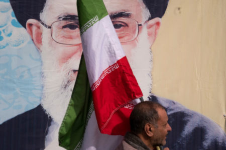 Imagem colorida de um cidadão do Irã carregando uma bandeira do país de frente a uma pintura do aiatolá Ali Khamenei - Metrópoles