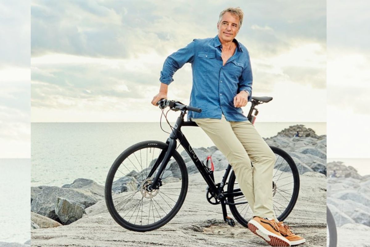 Foto colorida. Dan Buettner é um homem mais velho, branco e de cabelos grisalhos. Ela está perto de uma praia sentado em uma bicicleta - Metrópoles