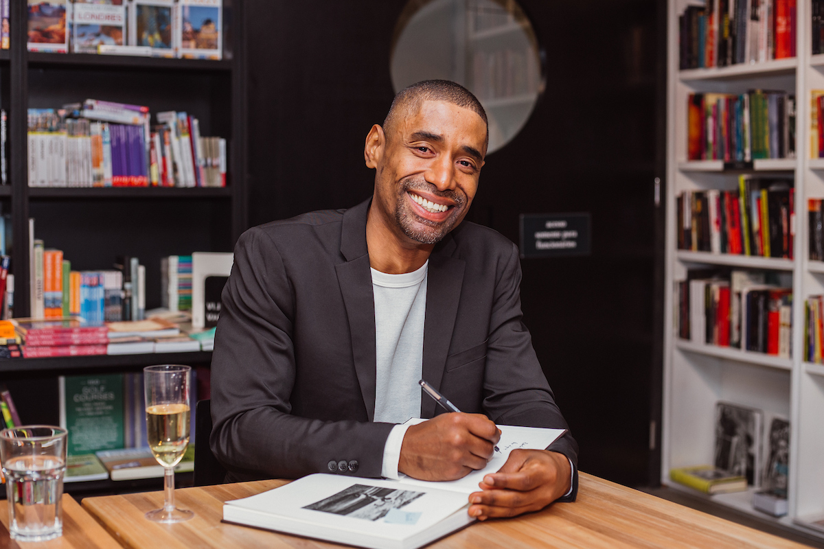 Foto colorida de Antonio Obá em uma livraria sentado em uma mesa e segurando um livro - Metrópoles