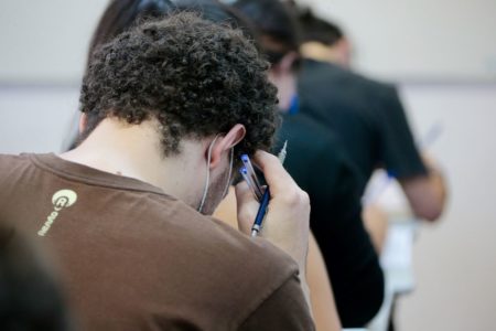 Imagem colorida mostra estudante branco com cabelo cacheado concentrado com a cabeça baixa durante prova da FUVEST. Ele veste uma camiseta marrom e segura duas canetas e uma lapiseira com a mão direita - Metrópoles