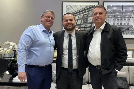 Imagem colorida mostra Tarcísio de Freitas, homem branco de camisa azul, Gil Diniz, de terno e gravata, e Jair Bolsoanro, de jaqueta preta e camisa branca. Os três estão abraçados, virados para a câmera, em uma sala de tons pasteis - Metrópoles