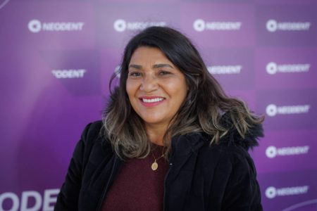 Imagem colorida de mulher vestida com casaco em fundo roxo enquanto sorri - Metrópoles