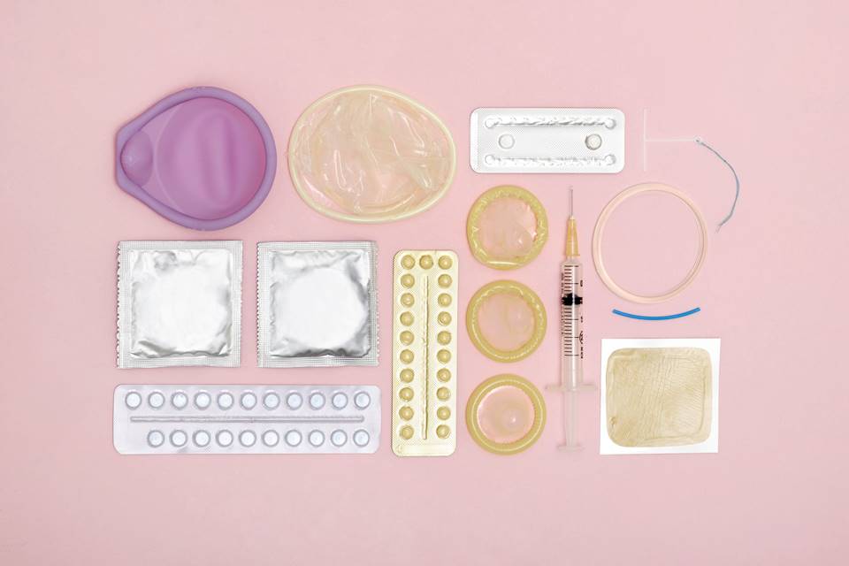 Foto de fundo rosa com objetos como camisinhas, pílulas e seringas espalhadas - Metrópoles