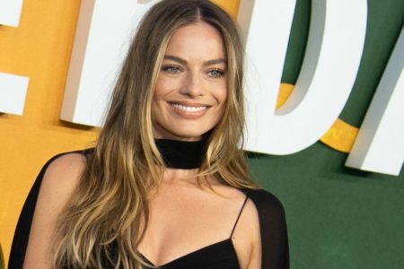 Foto colorida da atriz Margot Robbie sorrindo com um vestido preto e o cabelo solto - Metrópoles