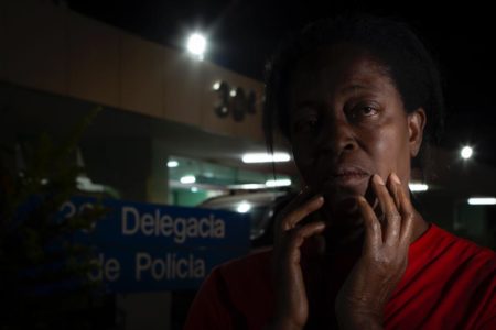 Imagem de mulher negra com camiseta vermelha em frente a delegacia com as mãos no rosto - Metrópoles