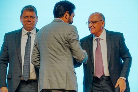 Imagem colorida mostra Geraldo Alckmin e Ricardo Nunes, ambos de terno e gravata, se cumprimentando, na frente de um fundo azul. A cena tem uma terceira pessoa, Tarcísio de Freitas, também de terno