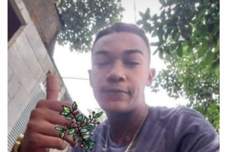 Foto colorida de Kauã Jazon da Silva em local aberto, com árvores ao fundo, fazendo "joinha", de camiseta lilás e corrente no pescoço