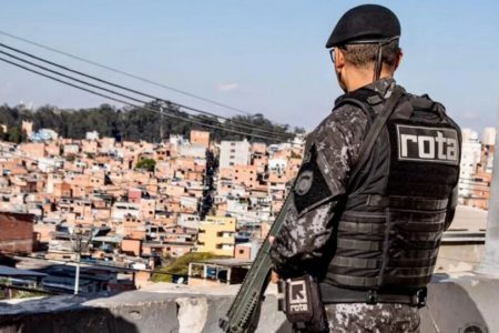 Em foto colorida policial da Rota, de costas, observa favela enquanto segura um fuzil - Metrópoles