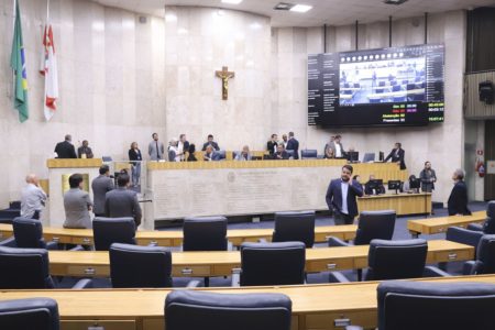 Imagem colorida mostra plenário da Câmara de SP, salão com cadeiras de couro azul e mesas com de madeira clara, com uma mesa diretora de mármore ao fundo e uma cruz cristã na parede. O salão tem alguns homens de terno em pé - metrópoles