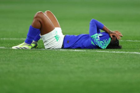 Mulher negra, vestida de uniforme azul, deitada em gramado de futebol com a mão sobre o rosto - Metrópoles