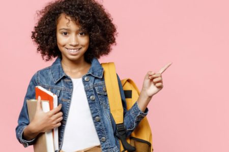 Menina negra de cabelos enrolados com livros nas mãos apontando para o lado com jaqueta jeans - Metrópoles