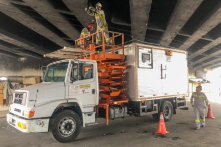 foto colorida, operários da Prefeitura trabalham na manutenção de viaduto. Eles estao num caminhão na parte de baixo do viaduto