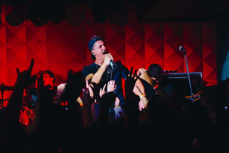 Imagem colorida mostra o cantor Gustavo Fildzz, da banda Aliados, cantando durante show da banda. Ele veste uma camiseta preta e um boné preto em um palco com fundo vermelho - Metrópoles