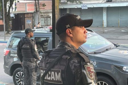 Dois policiais militares fardados e com colete à prova de balas; estão de pé, perto de viatura, em rua do Guarujá, foto colorida