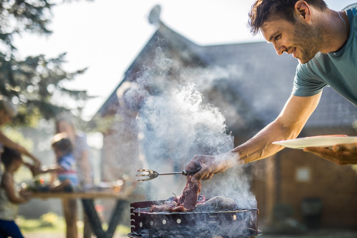 Foto colorida de homem branco colocando com a mão um churrasco na grelha. Fumaça sobe. Ele segura um garfo grande - Metrópoles