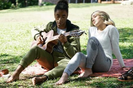 Imagem colorida de uma mulher loira sentada ao lado de um homem negro tocando violão, personagens do filme No Ritmo da Fé' - Metrópoles