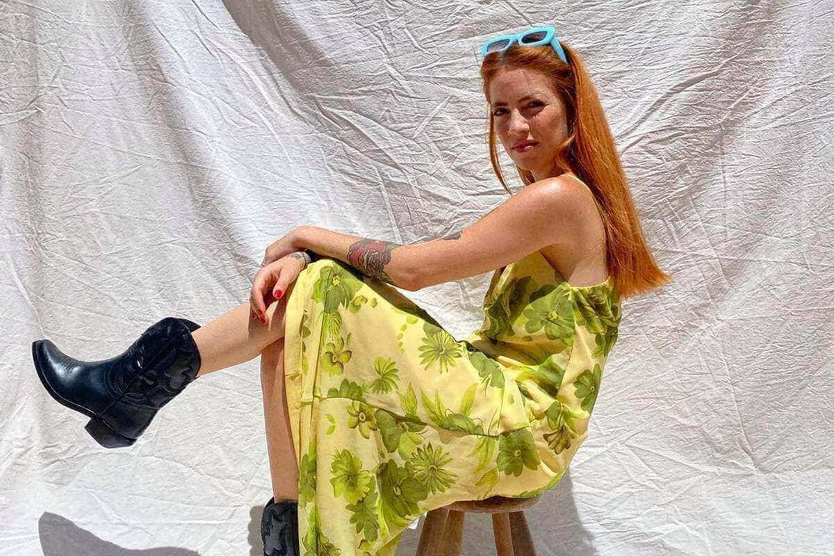 Na imagem com cor, mulher branca com os cabelos ruivos usa vestido estampado e botas pretas - Metrópoles