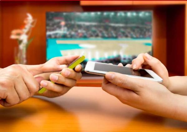 Fotografia colorida mostrando duas pessoas de lados opostos de uma mesa jogando nos celulares com uma TV ligada ao lado deles-Metrópoles