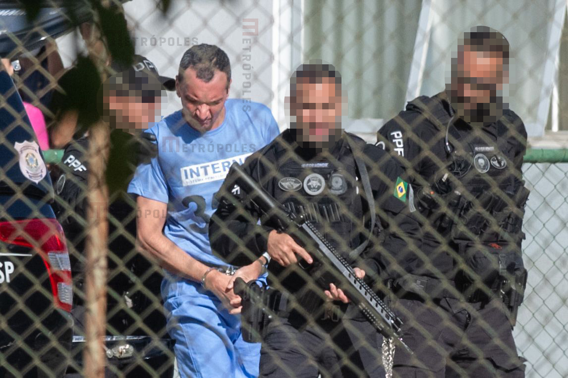 Marcola, homem branco, magro, de cabelos curtos e escuros, veste uniforme azul, usado na prisão; ele é escoltado por policiais de uniformes pretos e com rostos borrados