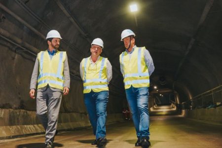 Imagem colorida mostra o prefeito Ricardo Nunes e mais dois homens andando em um túnel viário vazio. Eles vestem capacete branco e colete fluorescente - Metrópoles